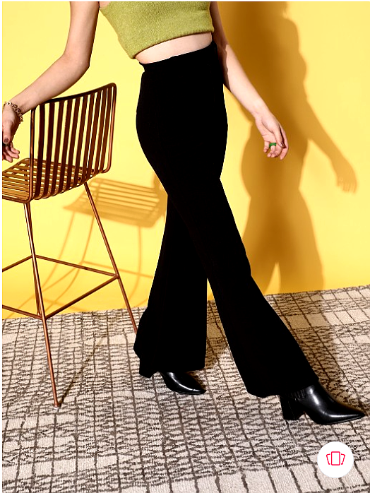 A woman modeling high-waisted black flared pants with a green crop top, captured from a side angle against a bright yellow backdrop.