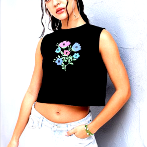 A young woman wearing a black sleeveless crop top with a colorful floral graphic print, paired with light blue denim jeans, posing confidently against a textured grey wall.