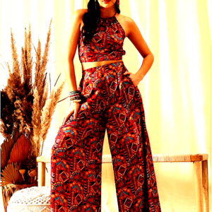 A woman wearing a vibrant red printed co-ord set, featuring a sleeveless halter neck crop top and high-waisted wide-leg trousers, posing confidently against a boho-themed backdrop.