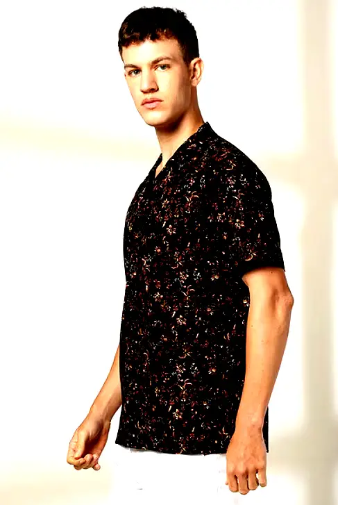 A male model wearing a black short-sleeve floral shirt with a relaxed fit and intricate multicolor floral patterns.