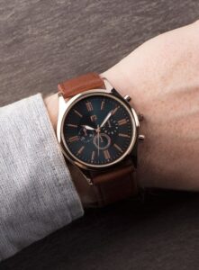 Close-up of a wristwatch with a brown leather strap, rose gold case, and a dark dial with rose gold details.