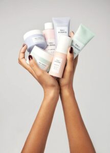 Two hands holding multiple pastel-colored skincare products from the Sali Hughes collection by Revolution, including jars and tubes in pastel shades.