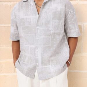 Person wearing a light grey short-sleeve button-up shirt standing against a light-colored brick wall.