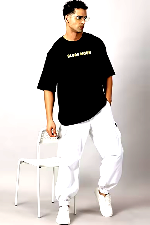 : A man wearing a black oversized T-shirt with "Blood Moon" text print paired with white cargo pants and sneakers, striking a casual pose beside a white chair.