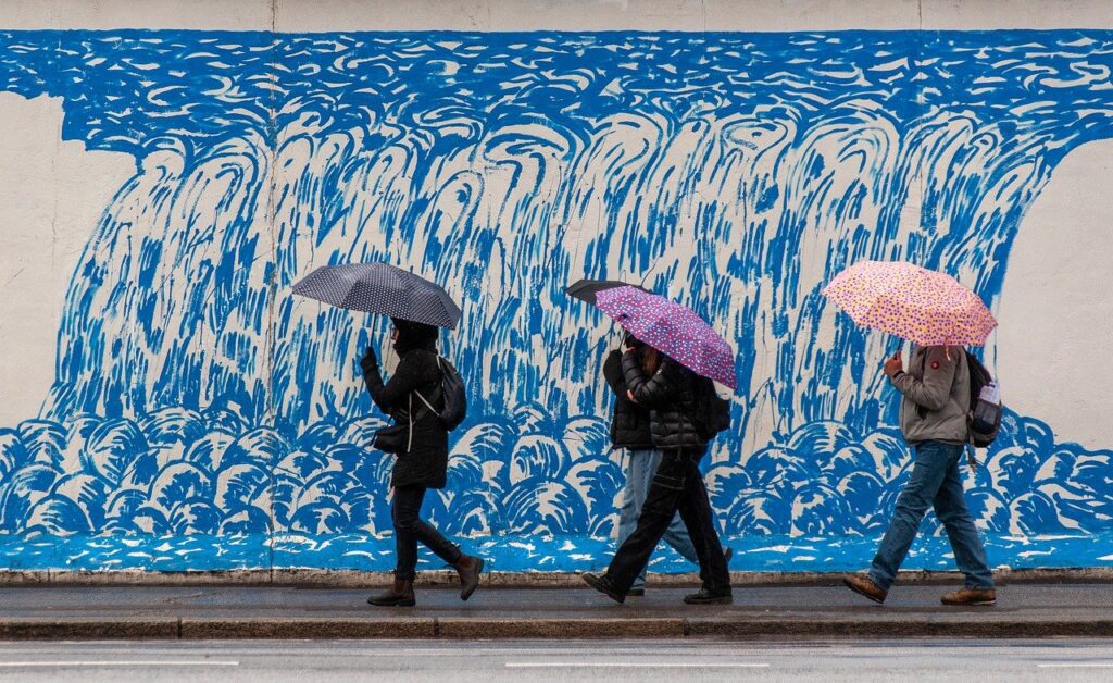 People walking under umbrellas past a blue wave mural on a rainy day.