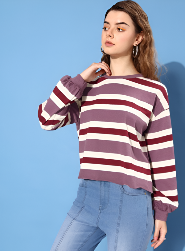 A fashionable young woman poses in a purple striped sweater and fitted denim jeans against a blue background, exuding a modern casual style.