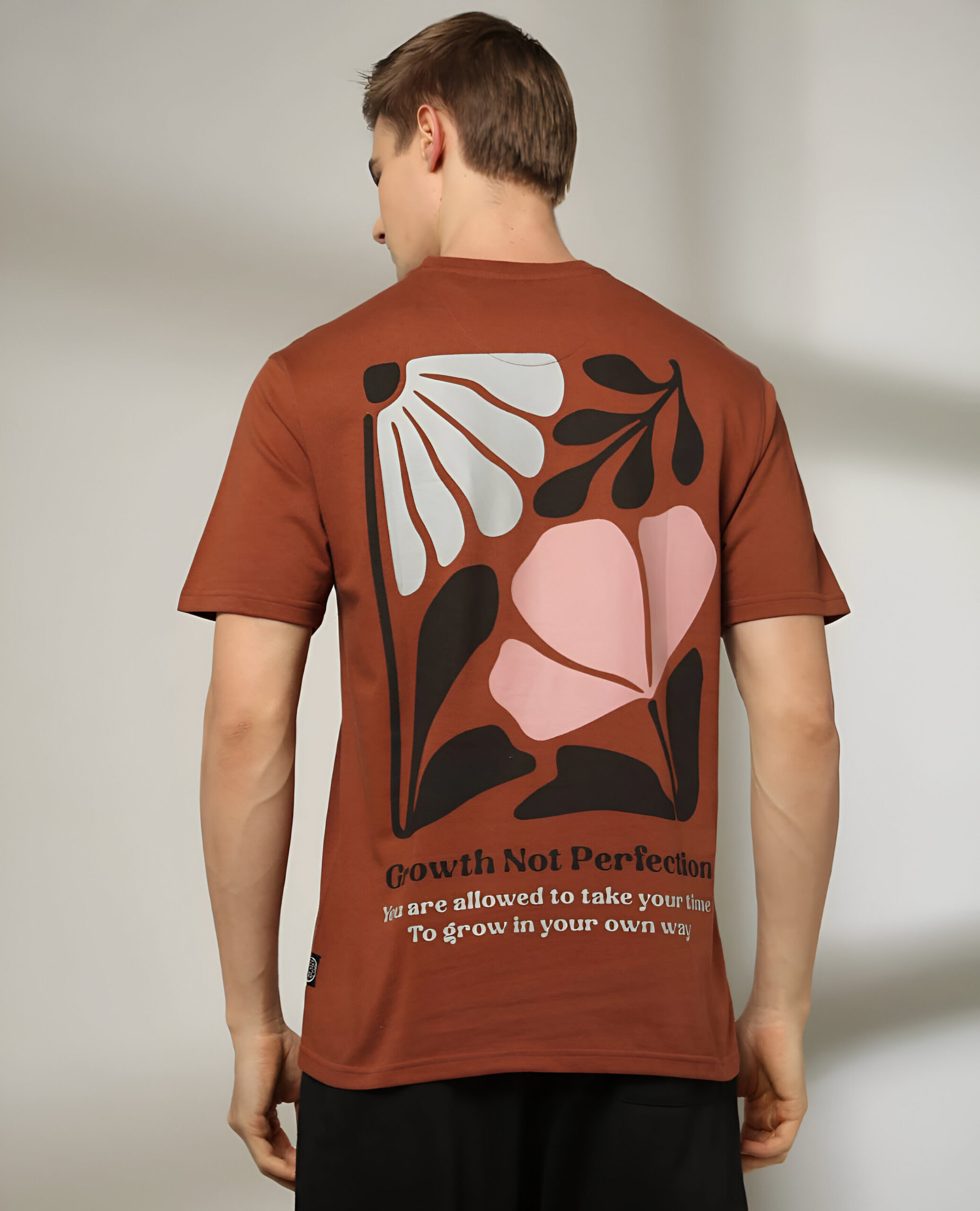 DNMX 3 "A man wearing a brown short-sleeved T-shirt with a large abstract floral print and the quote 'Growth Not Perfection' on the back, paired with black pants."