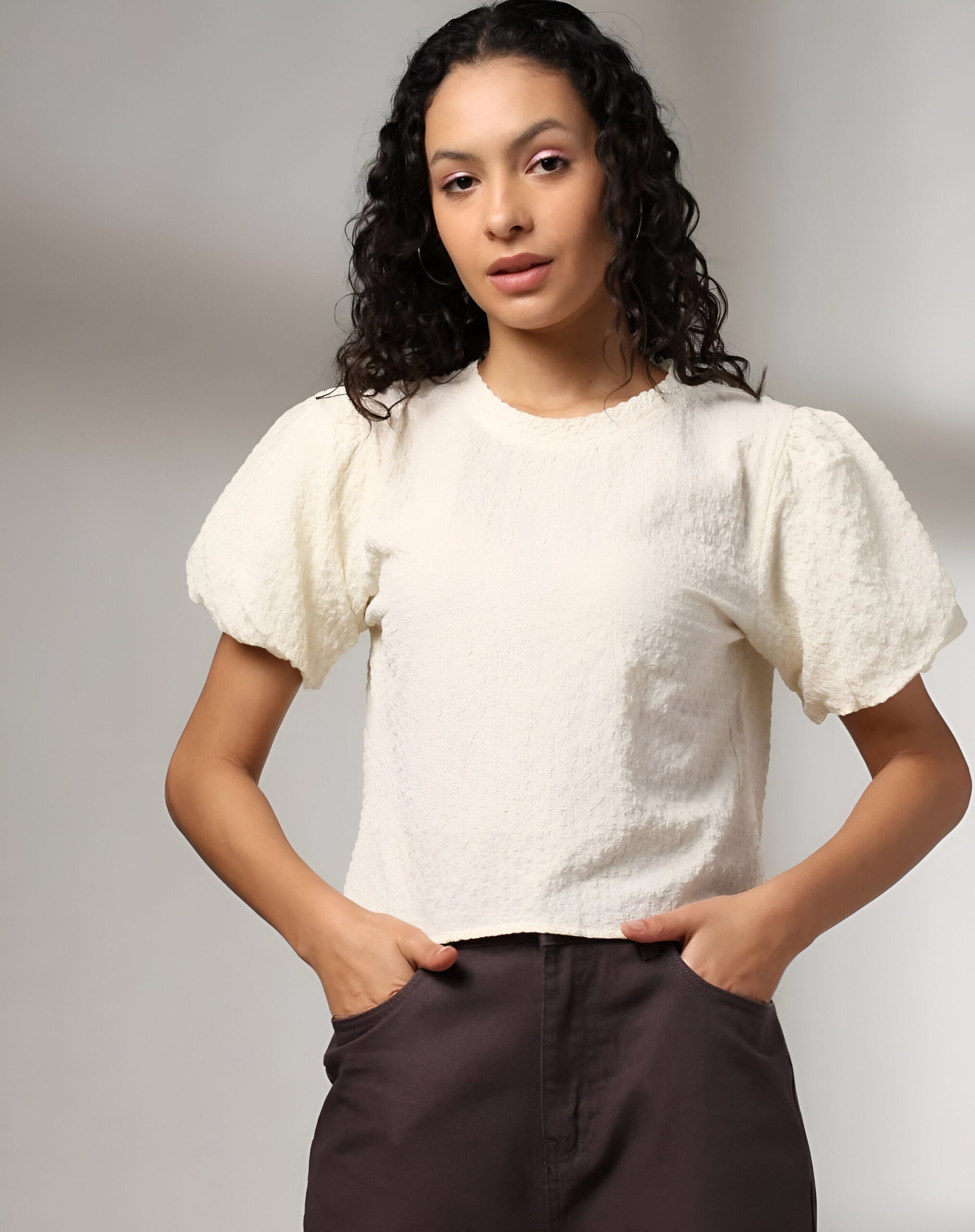 RIO 3 Woman wearing a white textured top with short puff sleeves, a round neckline, and high-waisted dark brown trousers, posing against a neutral background.