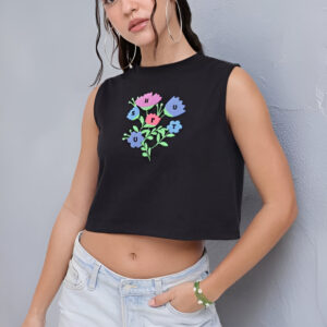 Woman wearing a sleeveless black crop top with colorful floral graphic print, light-wash high-waisted jeans, large hoop earrings, and a green bracelet, posing against a gray textured wall.