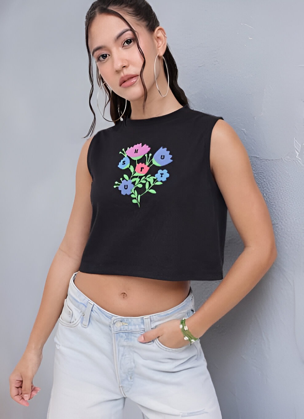 DressBerry 3 Woman wearing a sleeveless black crop top with colorful floral graphic print, light-wash high-waisted jeans, large hoop earrings, and a green bracelet, posing against a gray textured wall.