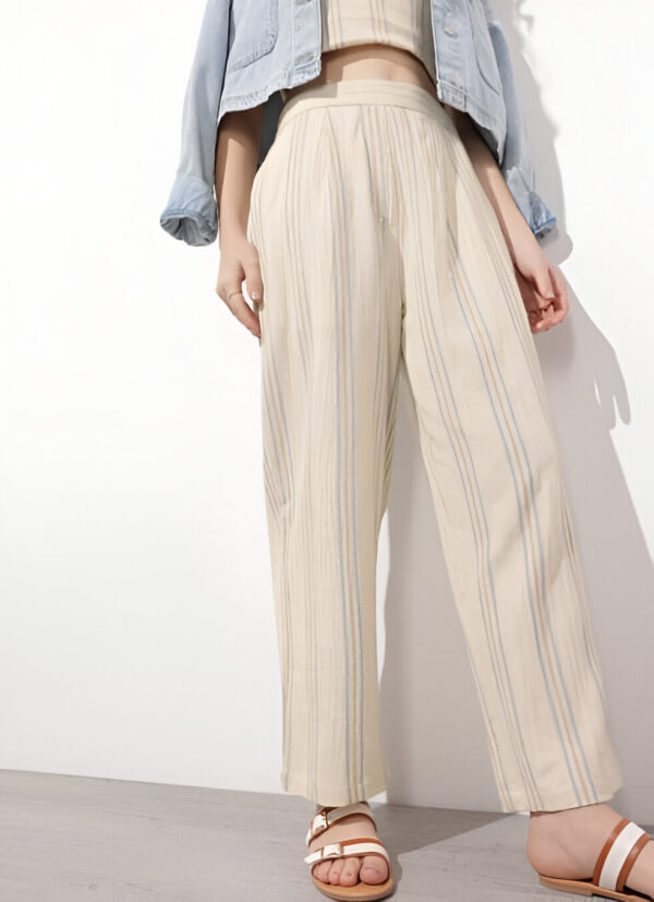 Woman wearing beige wide-leg pants with soft vertical stripes, paired with a cropped beige top, light-wash denim jacket, and brown strappy sandals, standing against a neutral background.