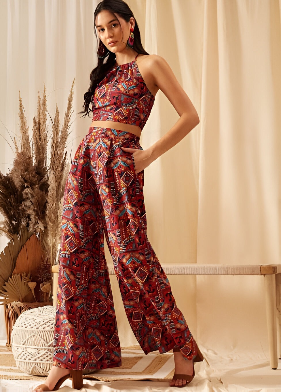  Woman wearing a sleeveless crop top and high-waisted wide-leg pants with a vibrant geometric print, paired with pink hoop earrings and open-toe heels, posing against a beige backdrop with pampas grass.