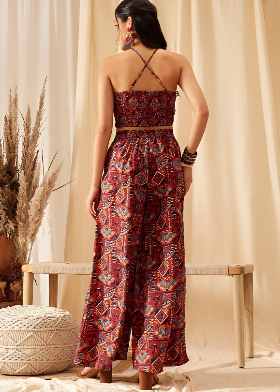 Back view of a woman wearing quiero two-piece bohemian outfit with a smocked crop top featuring crisscross straps and high-waisted wide-leg pants, accessorized with bold earrings and bangles, against a neutral backdrop with pampas grass and a wooden bench.
