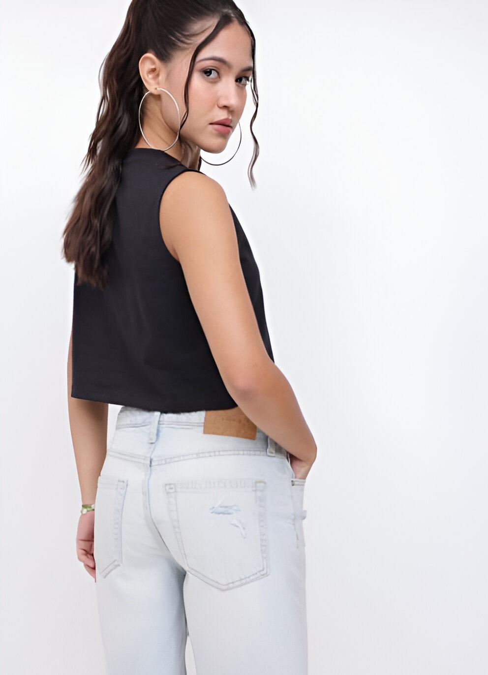DressBerry 4 Back view of a woman wearing a sleeveless black crop top and light-wash high-waisted denim jeans, accessorized with silver hoop earrings and a green bracelet, styled with a high ponytail.