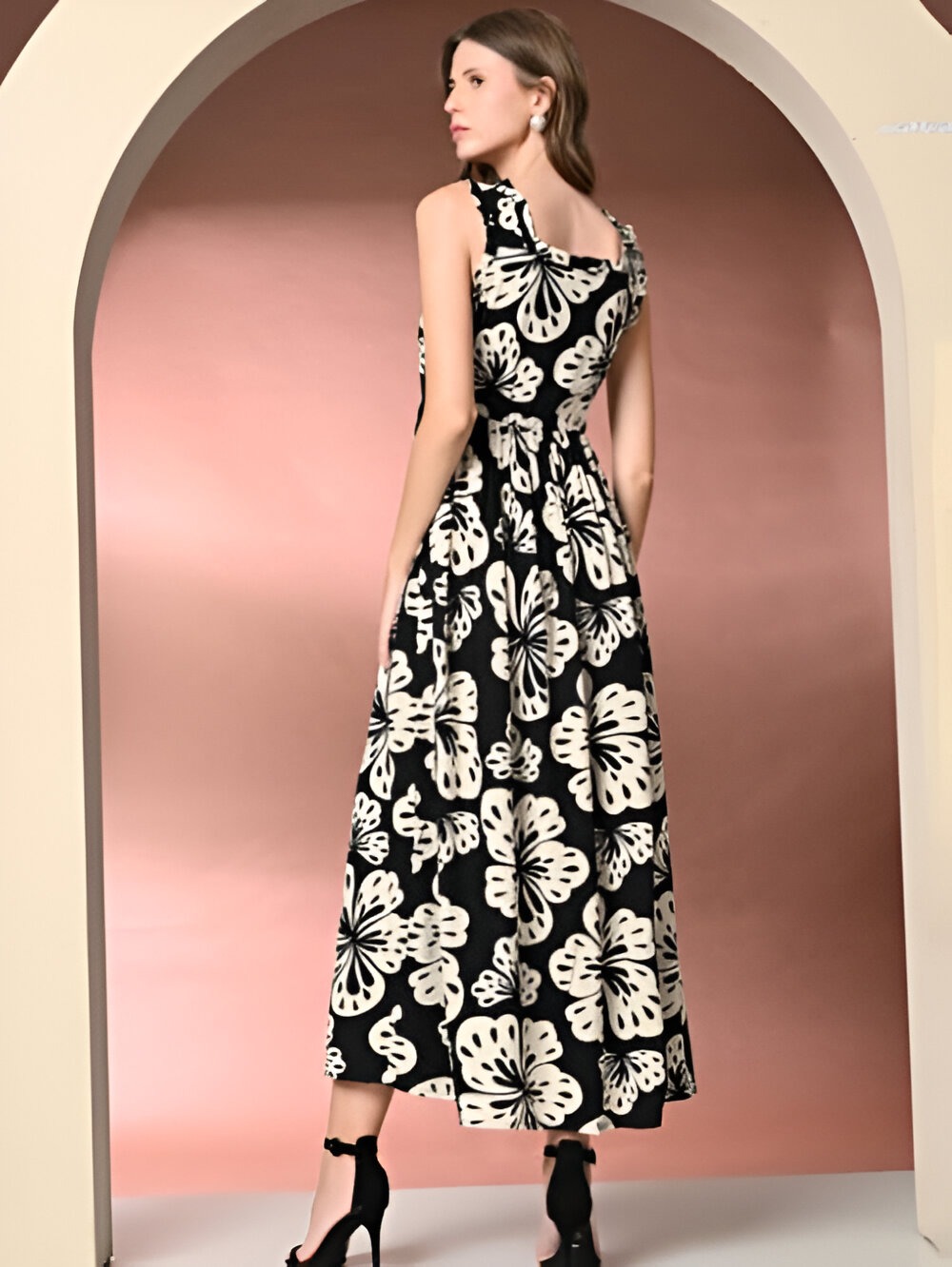 Back view of a woman wearing Daevish sleeveless black maxi dress with bold white floral patterns, a fitted waist, and paired with black high heels, posing under an arch with a peach-toned background.