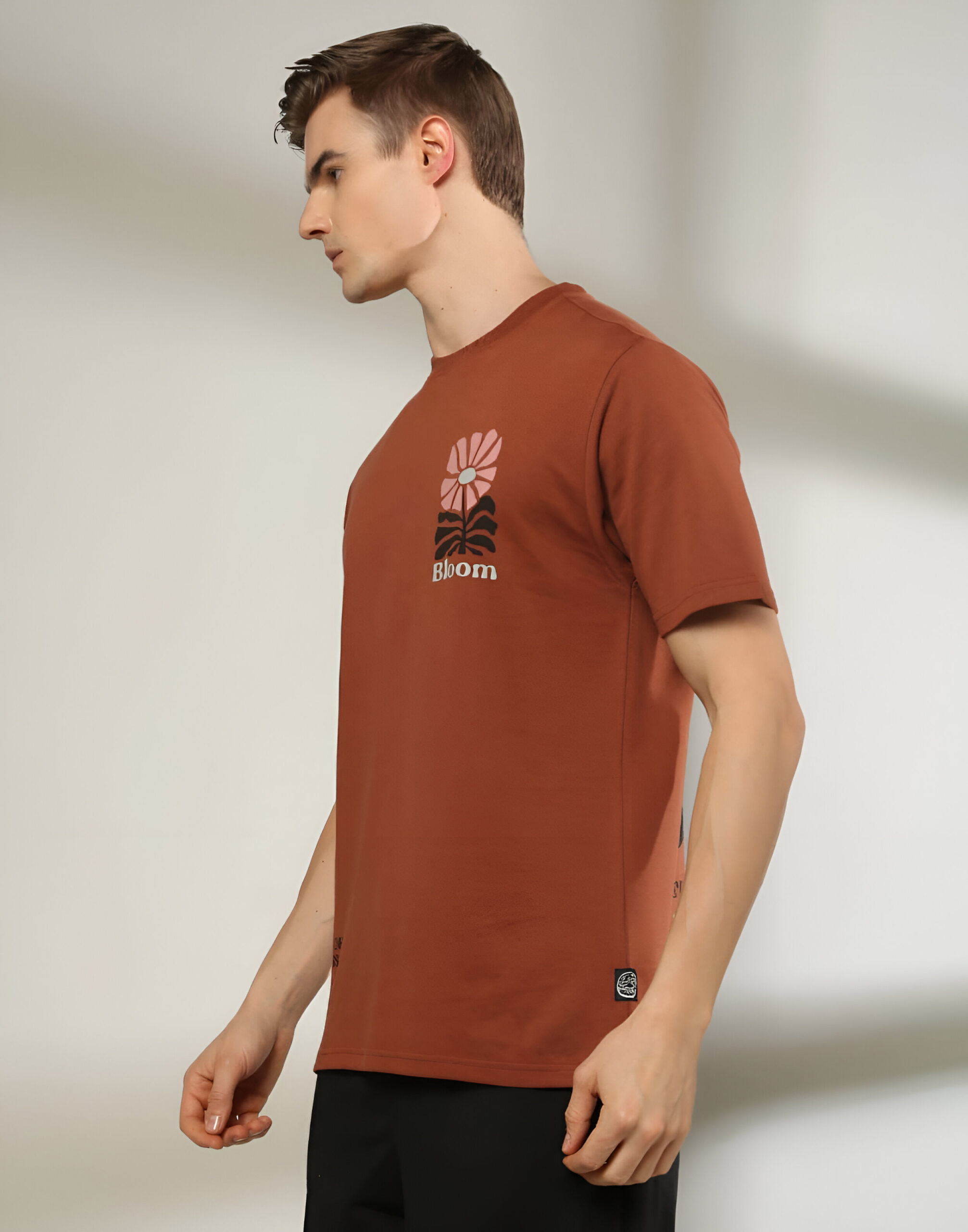 DNMX 4 "A man wearing a brown short-sleeved T-shirt featuring a small graphic of a pink flower and the word 'Bloom' on the chest, paired with black pants, standing against a neutral background."