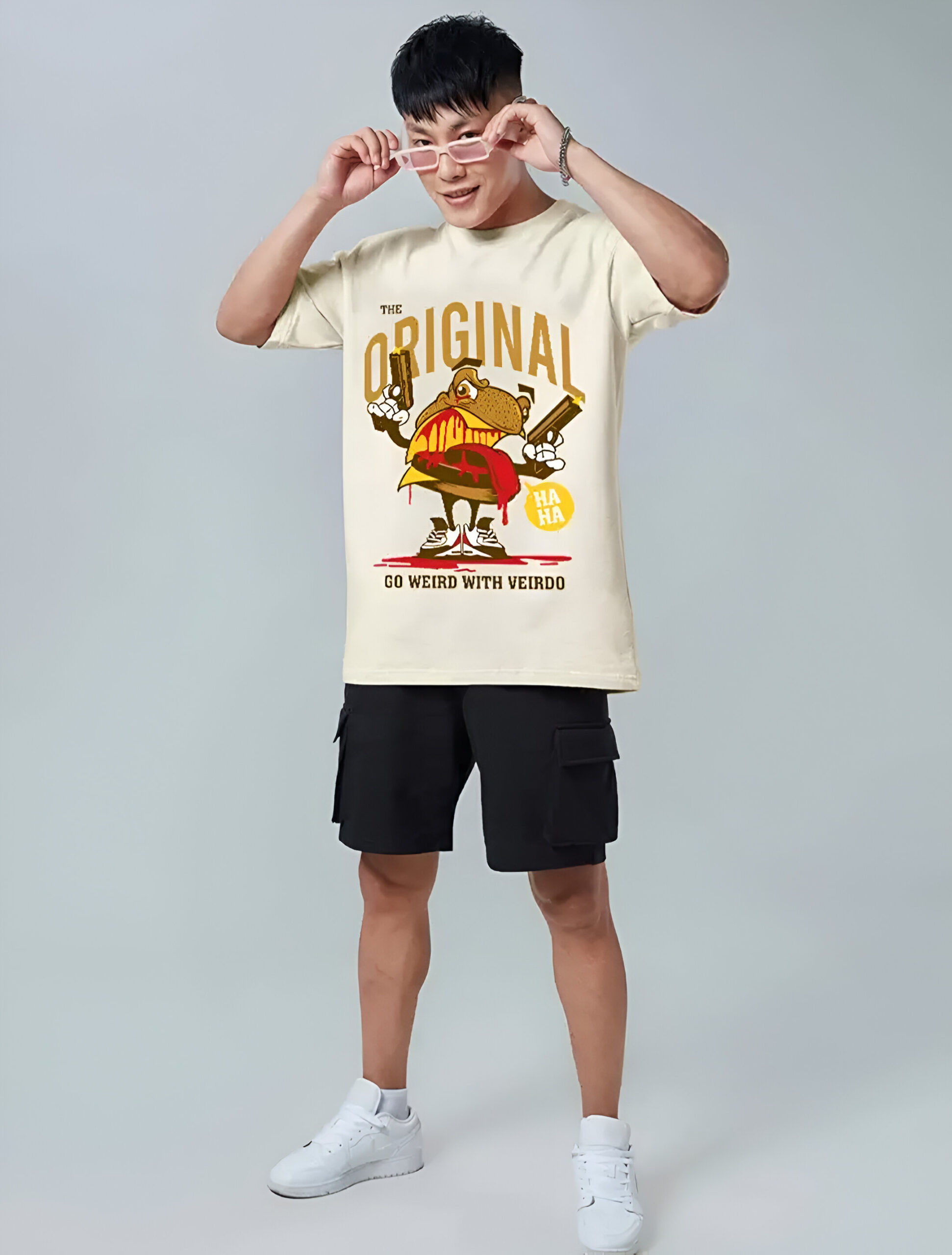 A young man wearing an oversized beige graphic T-shirt with a cartoon character, black cargo shorts, and white sneakers. He is adjusting his glasses while posing against a neutral gray background.