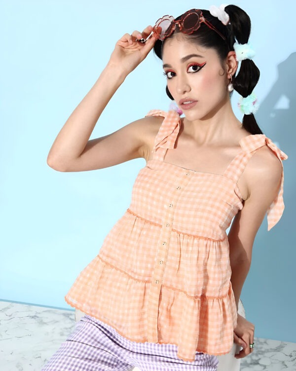 Woman wearing Quiero women peach gingham sleeveless tiered top with bow-tie straps, lilac gingham pants, oversized flower sunglasses, pearl earrings, and pastel scrunchies in her braids.