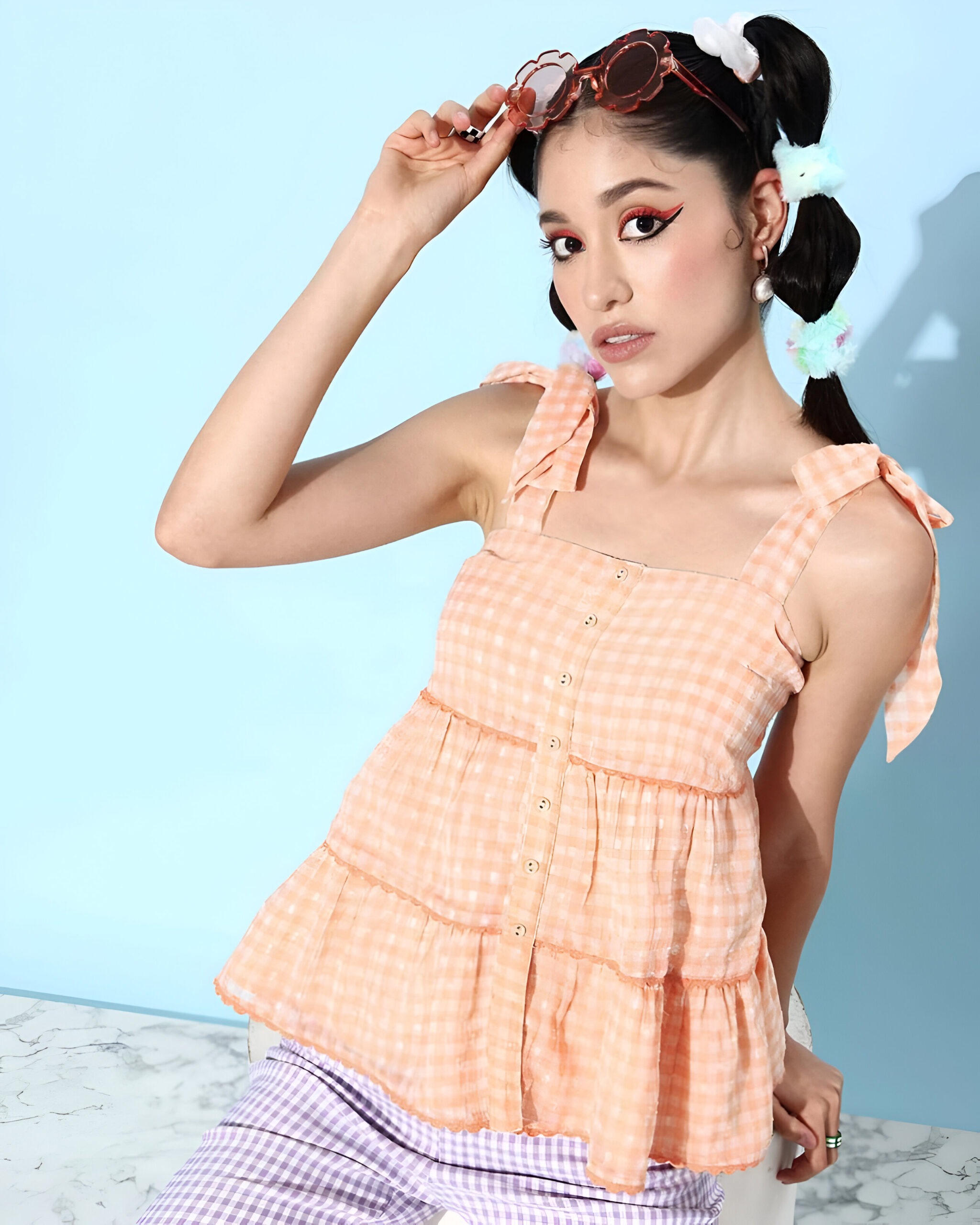 QUIERO 4 Woman wearing Quiero women peach gingham sleeveless tiered top with bow-tie straps, lilac gingham pants, oversized flower sunglasses, pearl earrings, and pastel scrunchies in her braids.
