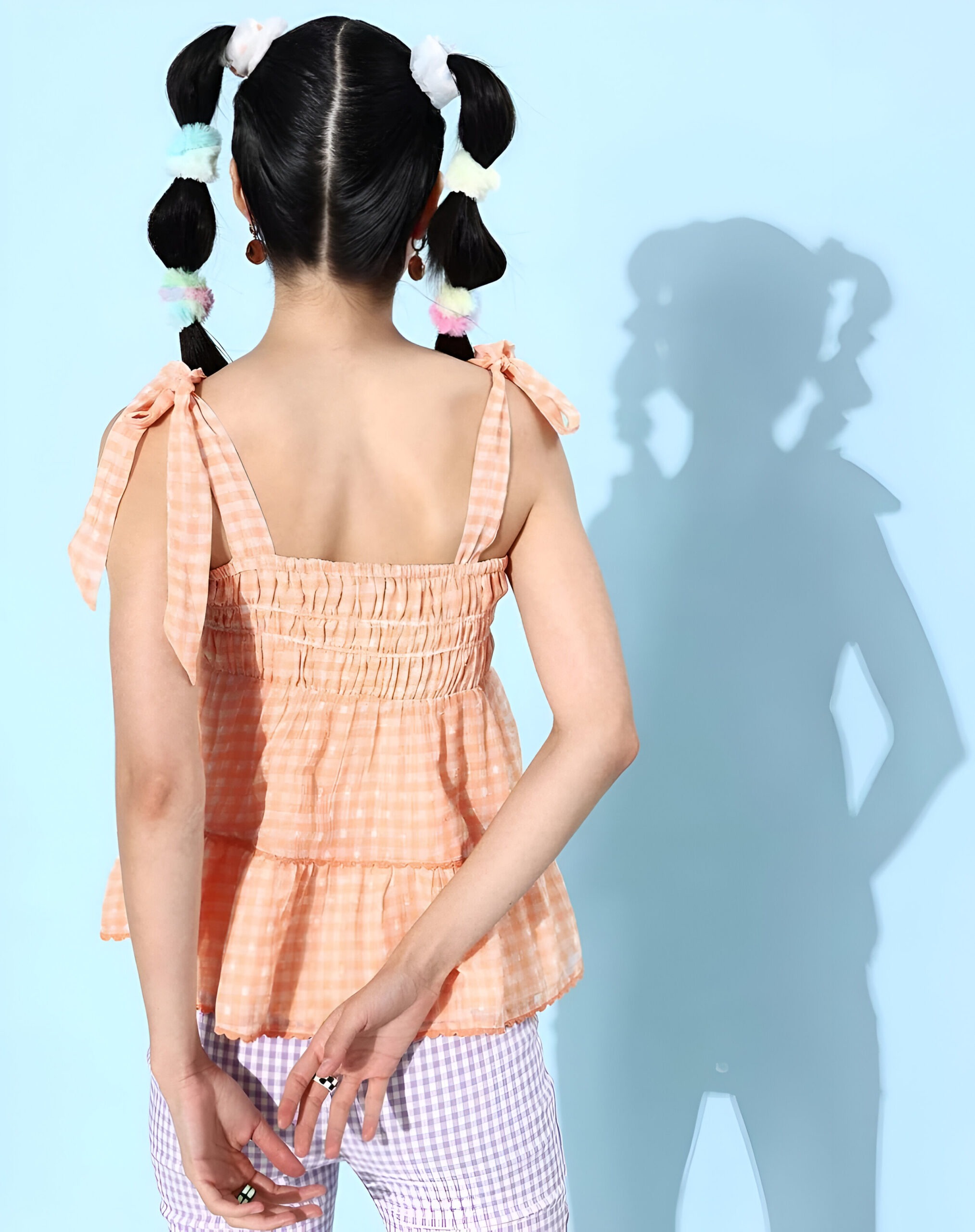 QUIERO 5 Back view of a woman wearing a peach gingham tiered top with bow-tie straps, smocked back, pastel scrunchie-adorned braids, and lilac gingham pants.