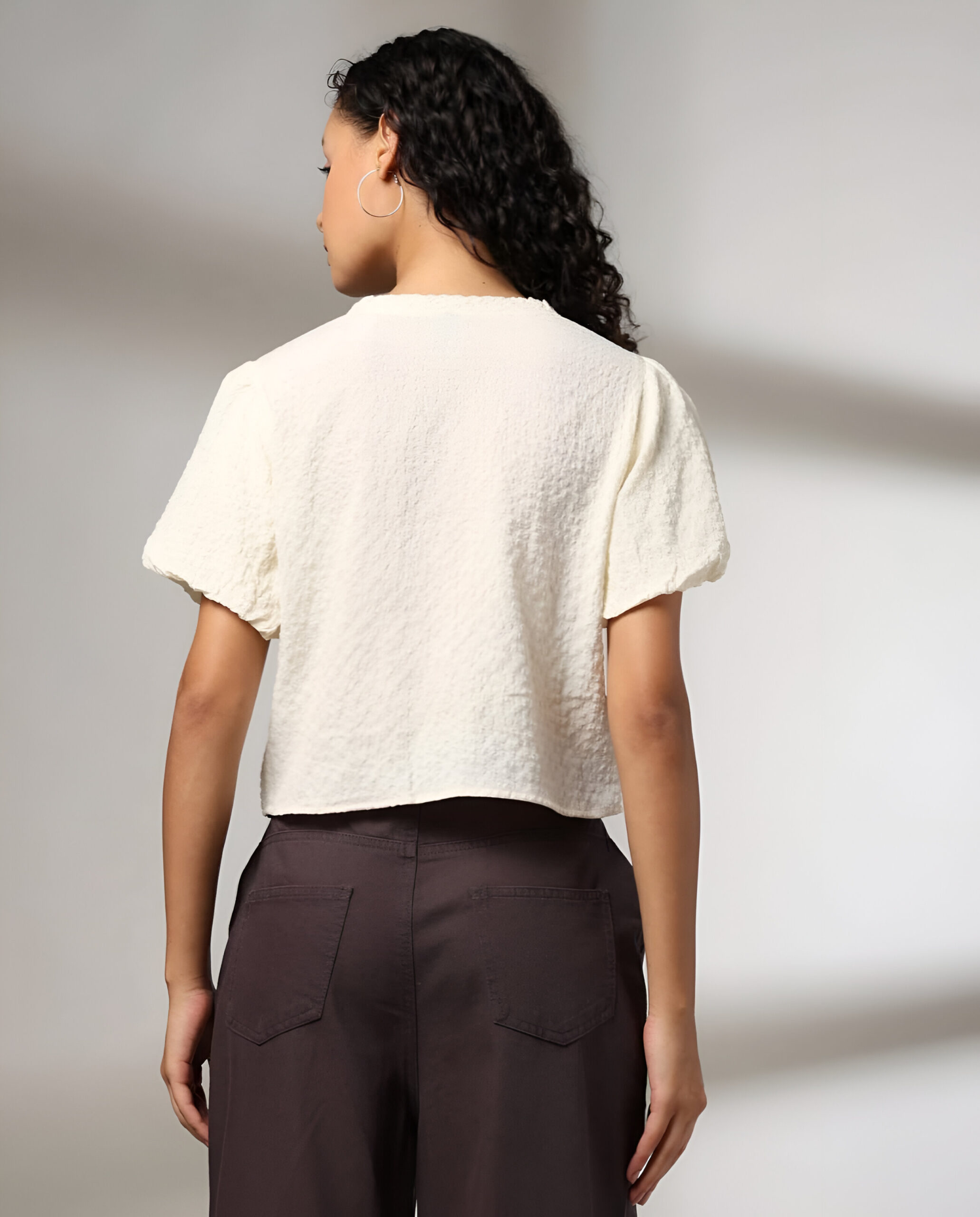 RIO 5 Back view of a woman wearing a white textured puff-sleeve top and high-waisted dark brown trousers, with curly hair and hoop earrings, against a neutral background.