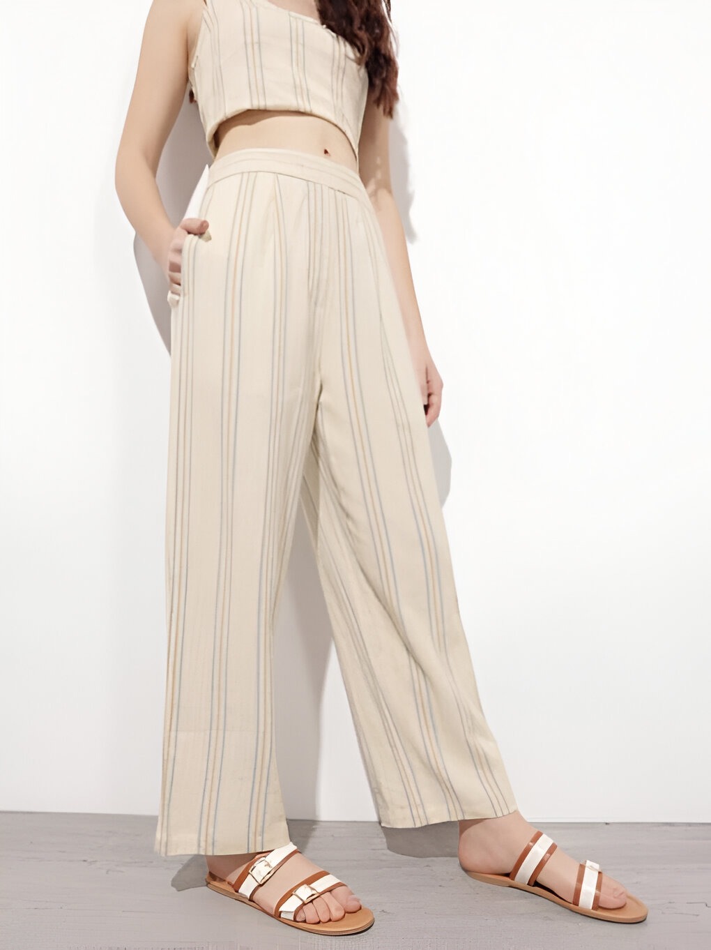 DressBerry 5 Woman wearing Dressberry beige wide-leg pants with soft blue and gray vertical stripes, a matching cropped top, and brown strappy sandals, posing against a neutral white background.