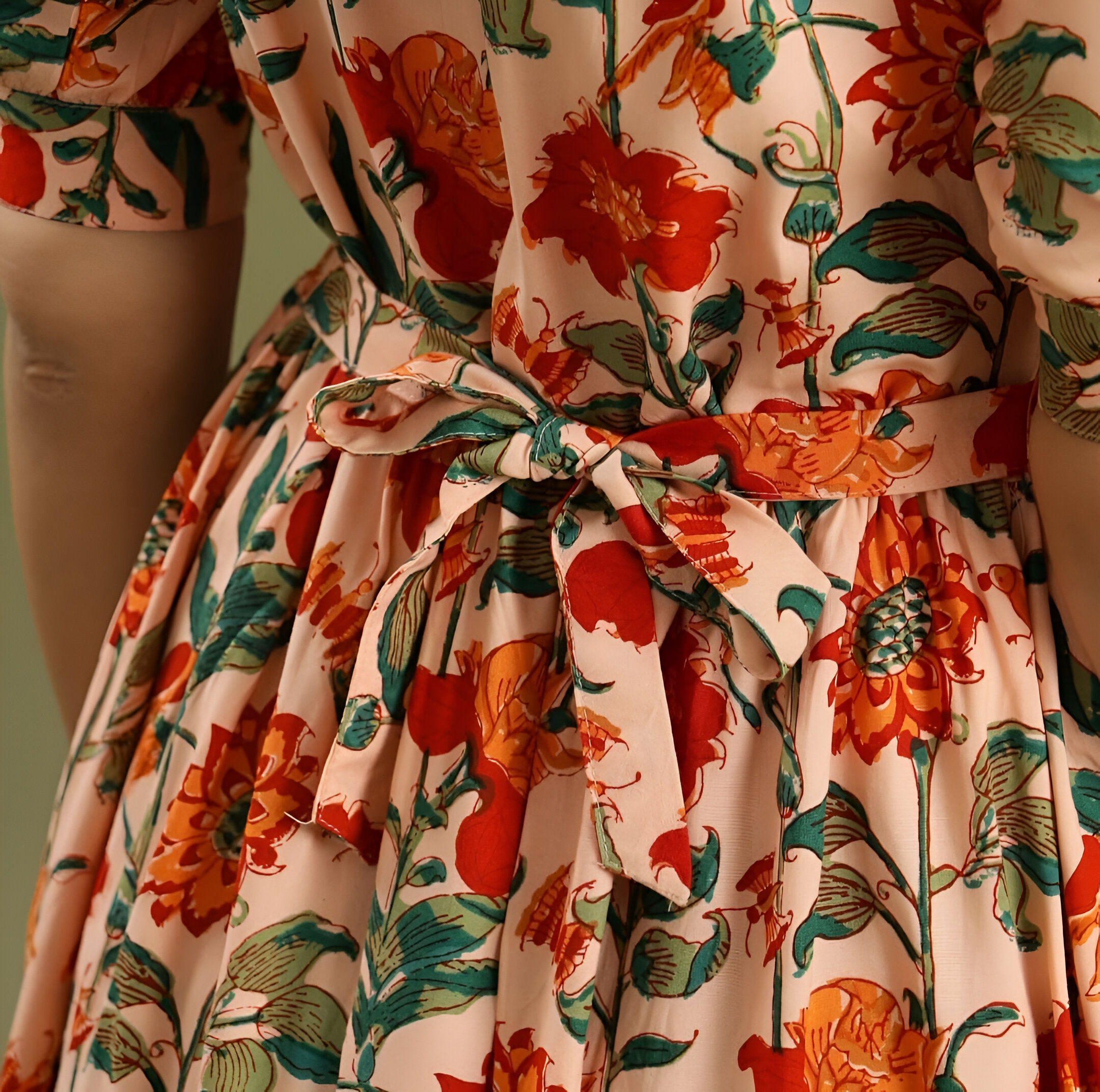 Negen 6 Close-up of a floral dress featuring a knotted waist tie, vibrant red and orange flowers, green leaves, and soft pleats on beige fabric.