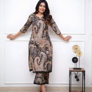 Woman wearing a paisley-printed black kurta set with gold and beige patterns, paired with matching trousers, black heels, and maroon jhumka earrings, posing against a white background.