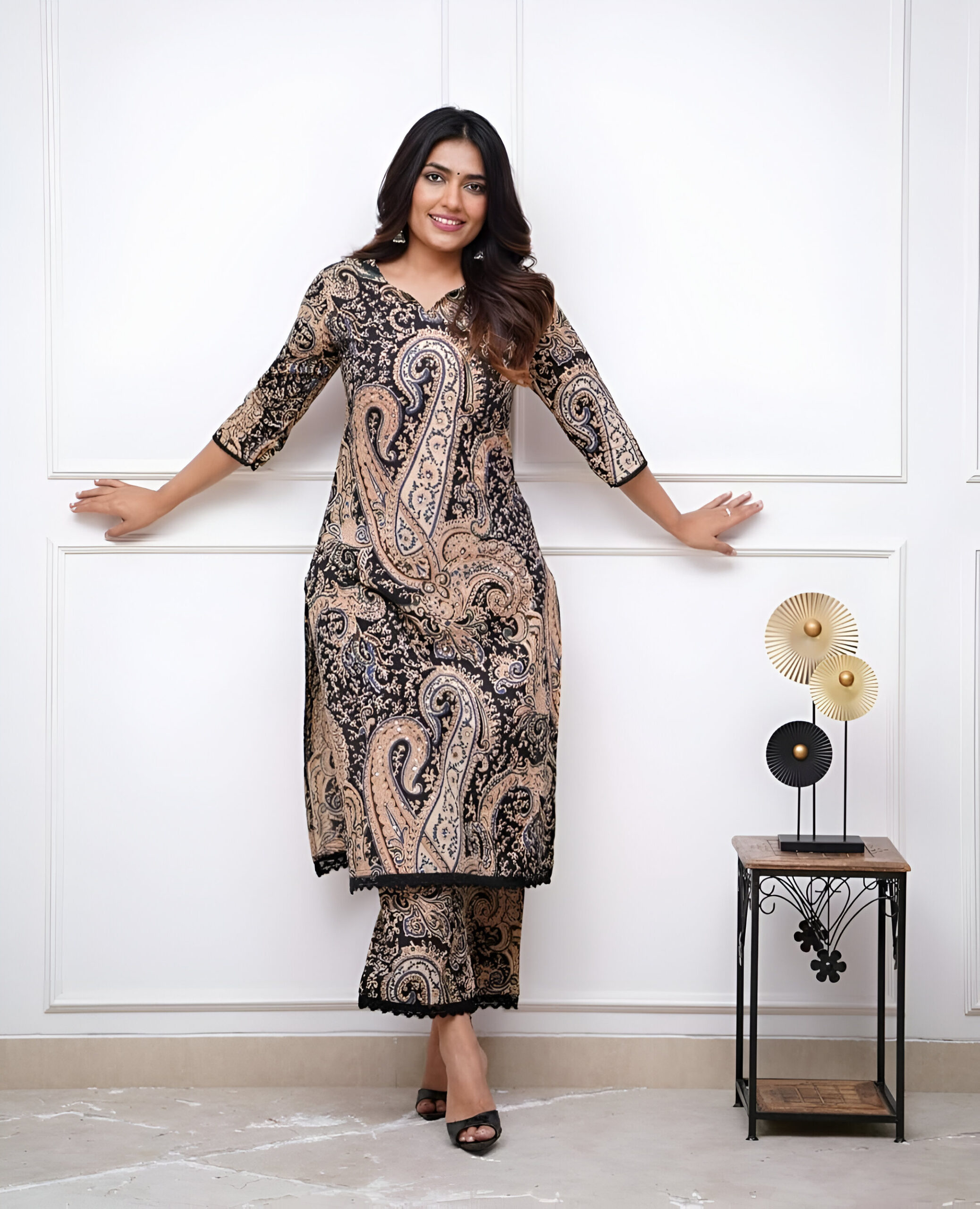 Nermosa 5 Woman wearing a paisley-printed black kurta set with gold and beige patterns, paired with matching trousers, black heels, and maroon jhumka earrings, posing against a white background.