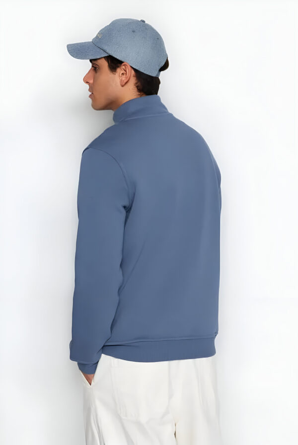 Back view of a man in a blue quarter-zip pullover, blue cap, and light trousers, standing against a plain white background.