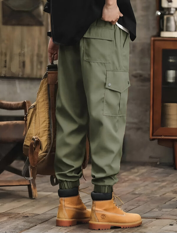 Back view of a person wearing olive green cargo pants with side and back pockets, tan work boots, and holding a brown backpack, standing in a room with wooden floors and vintage decor.