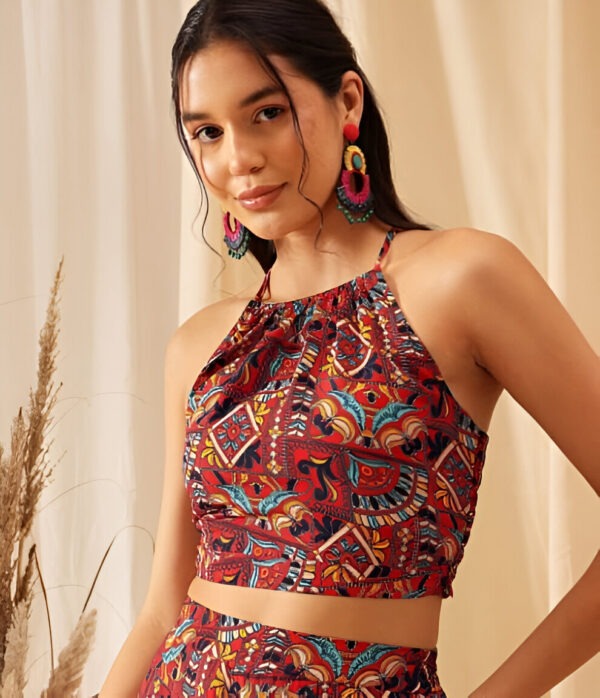 Woman wearing a sleeveless bohemian crop top with a halter neckline, featuring vibrant geometric patterns in red, blue, and orange, accessorized with oversized multicolored hoop earrings, against a beige backdrop with pampas grass.