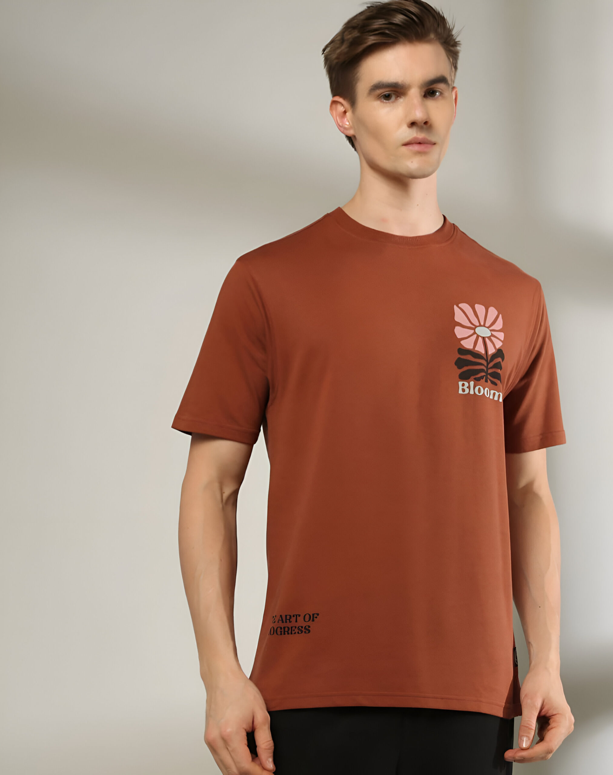 DNMX 5 "A man wearing a brown short-sleeved T-shirt featuring a small flower graphic with the word 'Bloom' on the chest and 'Part of Progress' text near the bottom, paired with black pants, standing against a neutral background."