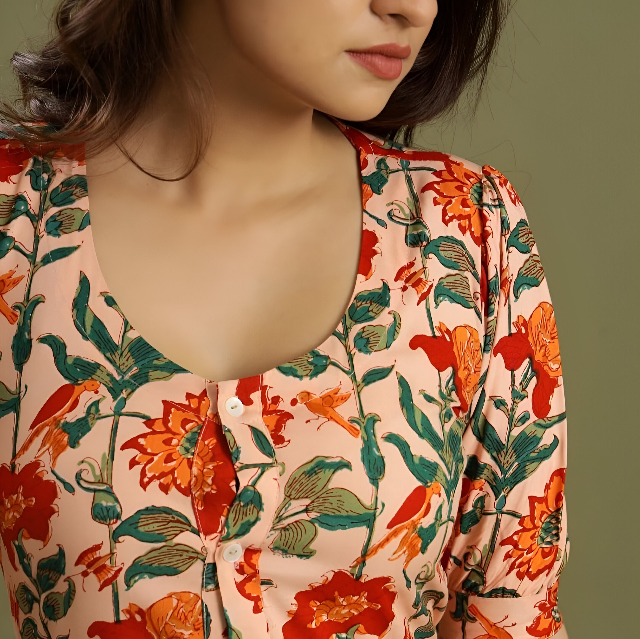 Negen 7 Close-up of a floral dress neckline showcasing red and orange flowers, green leaves, white buttons, and a round neckline on beige fabric.