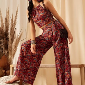 Woman wearing a sleeveless crop top and wide-leg pants with a vibrant geometric print, accessorized with multicolored hoop earrings, stacked bangles, round sunglasses, and a woven crossbody bag, posing against a beige backdrop with pampas grass and a wooden bench.