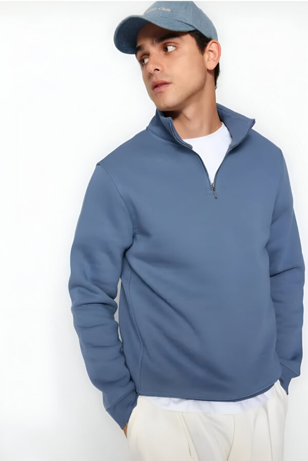 A man dressed in a blue quarter-zip pullover, white undershirt, and cap, standing against a plain white background.