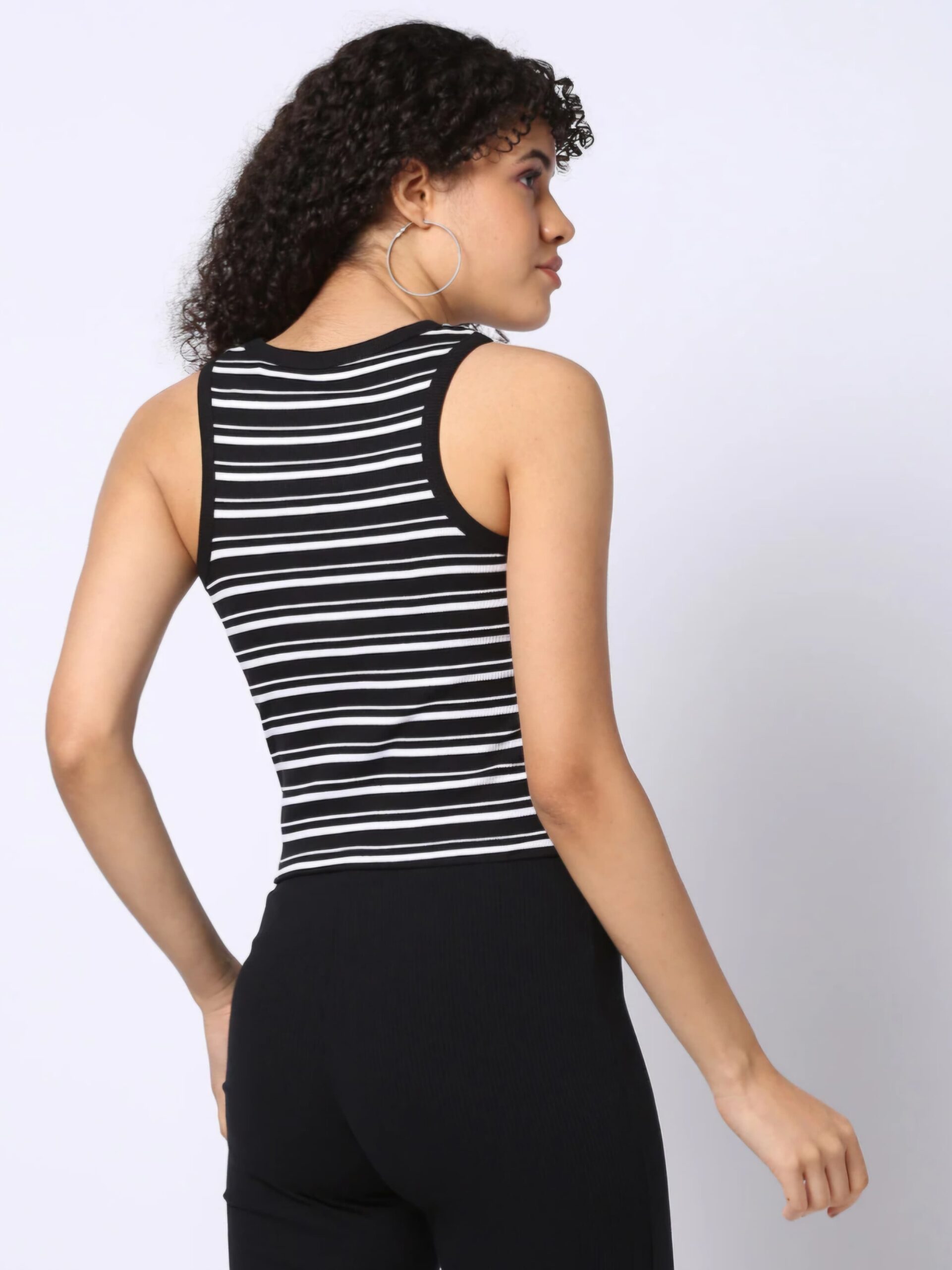 RIO BASIC 4 Here is a suggested set of metadata for the image you provided: Title: Striped Racerback Tank Top for Women Text Description: A woman wearing a black and white striped racerback tank top paired with black ribbed pants. She has curly hair, large hoop earrings, and is looking to the side against a neutral white background. Caption: Black and white striped racerback tank top styled with black ribbed pants and hoop earrings. Alternative Text: A woman wearing a black and white striped crop tees for women, looking to the side with hoop earrings and curly hair.