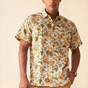 "A man wearing a short-sleeved floral print shirt with yellow, green, and brown flower patterns on an off-white background, paired with light brown shorts, a wristwatch, and silver rings, standing against a beige wall."