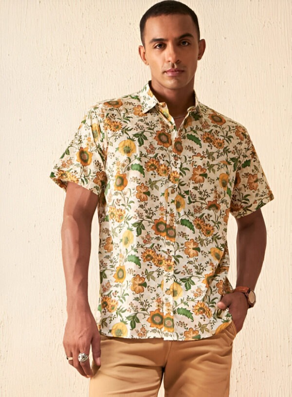 "A man wearing a short-sleeved floral print shirt with yellow, green, and brown flower patterns on an off-white background, paired with light brown shorts, a wristwatch, and silver rings, standing against a beige wall."