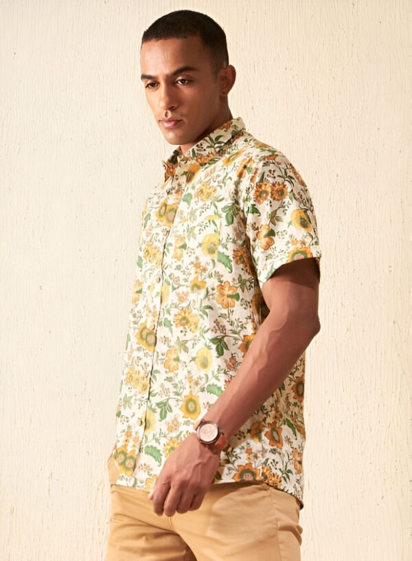 A man is shown in a side profile pose against a textured beige background. He is wearing a short-sleeved button-down floral shirt featuring a vibrant pattern of yellow flowers, green leaves, and brown accents on an off-white base. The shirt fits well, showcasing a relaxed yet polished style. He pairs the shirt with light brown shorts and accessorizes with a classic wristwatch on his left wrist. His confident posture and focused expression enhance the summery vibe of the outfit.