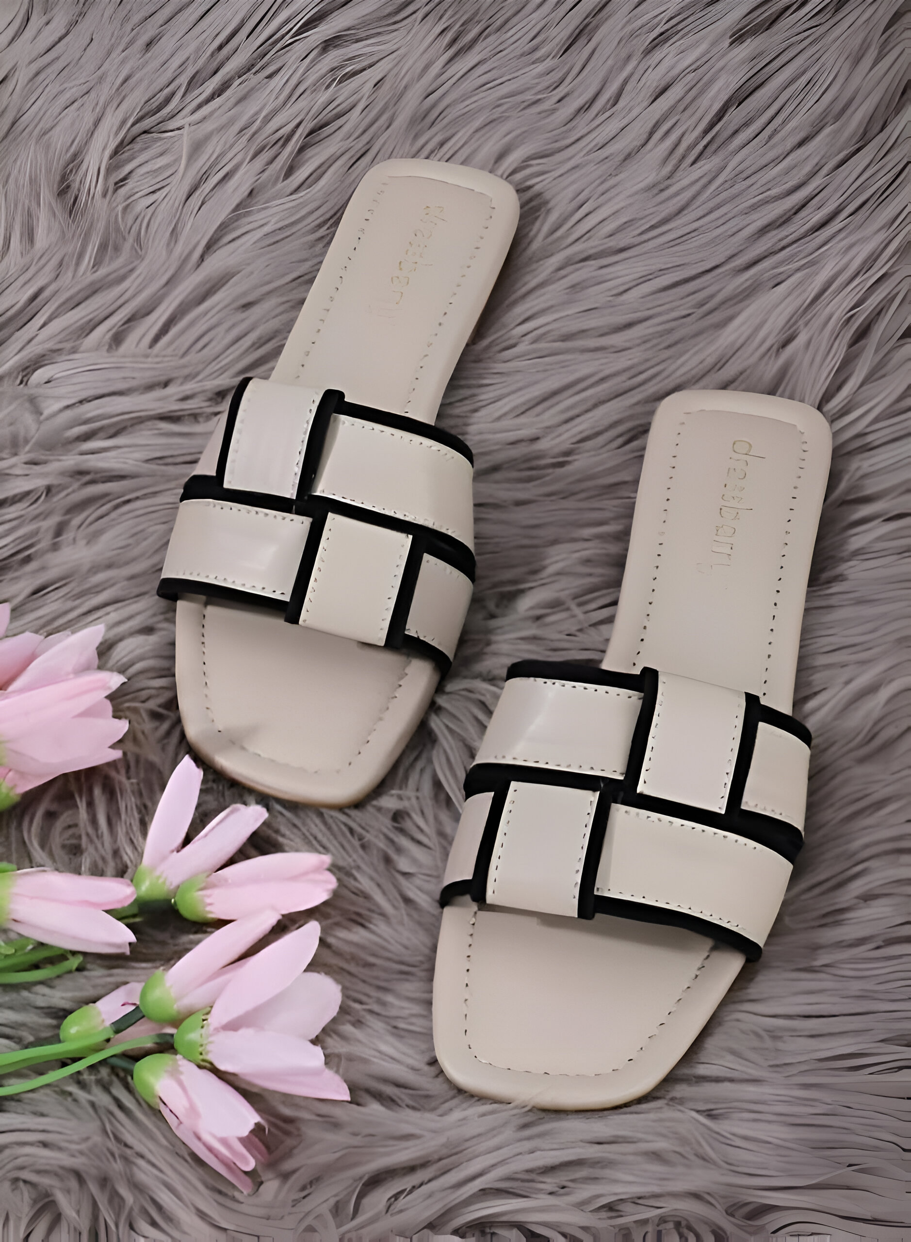 Dressberry (Women) 4 A pair of dressberry footwear beige slip-on sandals with black piping accents, displayed on a gray fur background with pink flowers beside them.